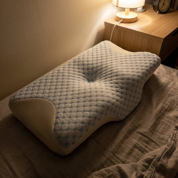 The Serenium Relief Pillow on a bed beside a lit bedside lamp